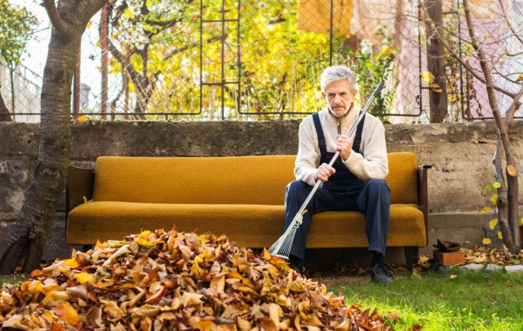 A senior man sitting in his backyard with a broomstick after getting tired of gathering dry leaves.